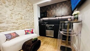 A modern living room with a stone wall, a white sofa with colorful cushions, a wooden coffee table, and a kitchen area with black cabinets and a glass coffee table. - Paris Marais by Melvane (Paris)