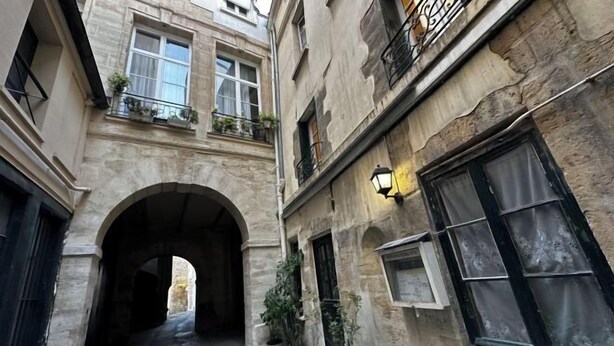 A narrow alleyway with a stone archway, potted plants, and a building with a balcony and a lit wall-mounted lamp. - Paris Marais by Melvane (Paris)