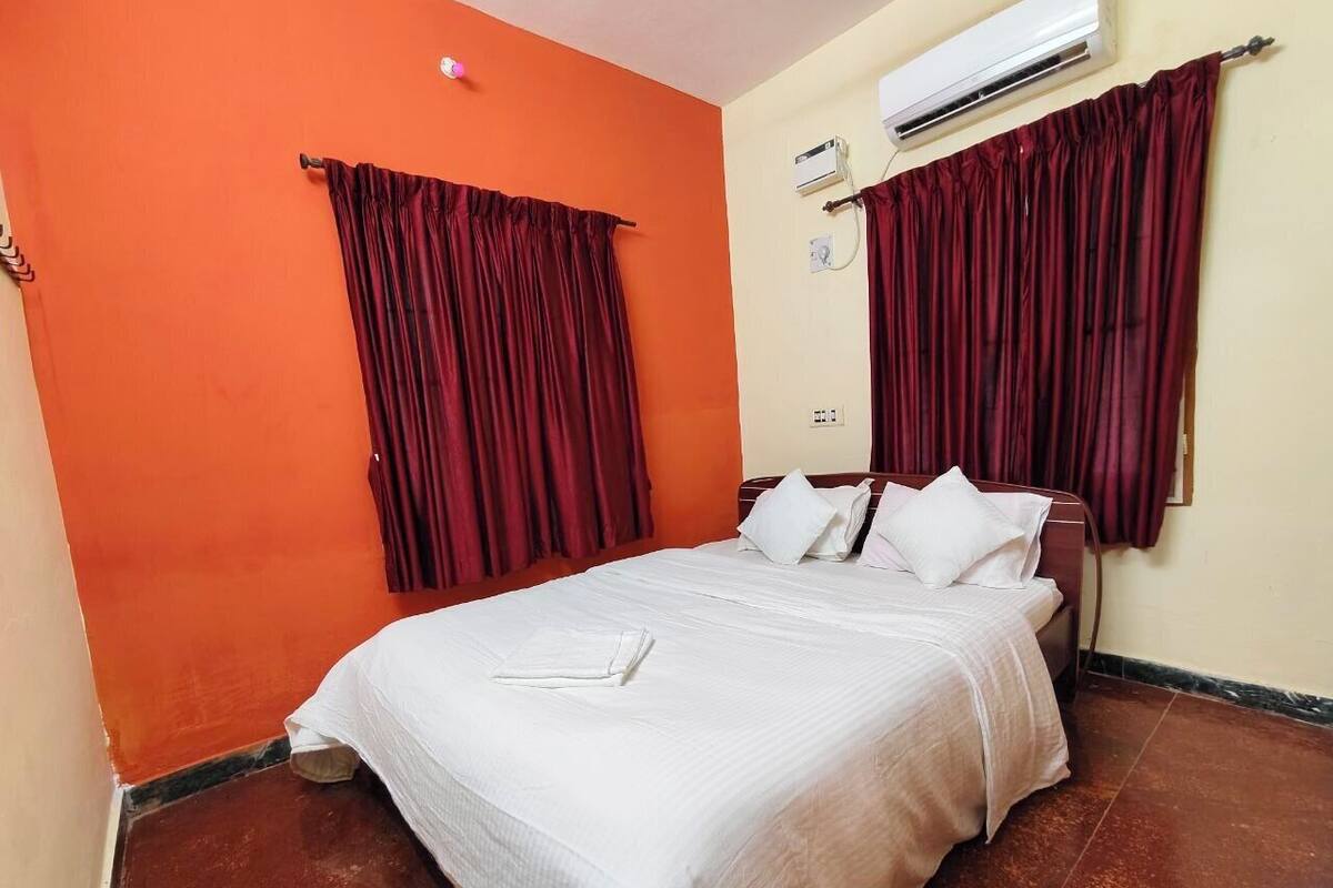 A hotel room with a bed, two red curtains, an air conditioning unit, and a white pillow on the bed.