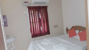 Un dormitori amb llit, una taula de nit, aire condicionat fixat a la paret, una finestra amb cortines i una presa elèctrica.