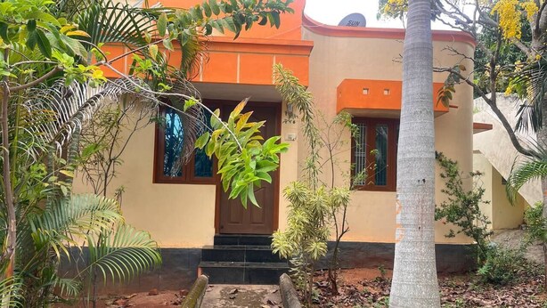 Edifici de dues plantes amb una porta marró i finestres, envoltat de vegetació i un petit camí que condueix a l'entrada.
