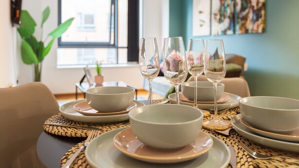 A dining table set with bowls and wine glasses, a potted plant, and a window with a view of buildings. - Modern 2 Bed apartment in Liverpool, City Centre (Merseyside)