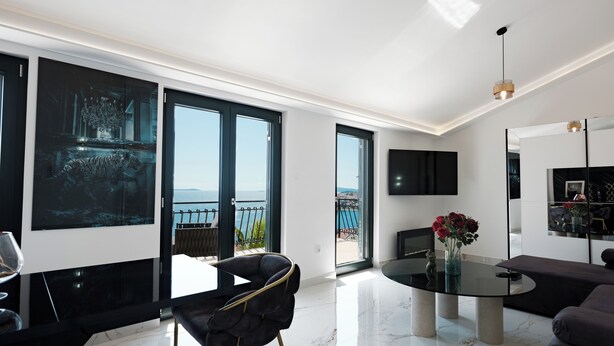 A modern living room with a large window, a round glass table, a black sofa, and a black chair. - Villa Aea Del Mare: Luxury 5BR Villa with Pool, Wellness & Sea Views - Sleeps 12 (Okrug Gornji)