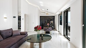 A modern living room with a glass coffee table, a dark sofa, and a dining area with a marble backsplash. - Villa Aea Del Mare: Luxury 5BR Villa with Pool, Wellness & Sea Views - Sleeps 12 (Okrug Gornji)