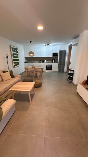 Una sala moderna con sofá, una mesa de centro y un comedor. Se ve una cocina con armarios y electrodomésticos blancos al fondo.