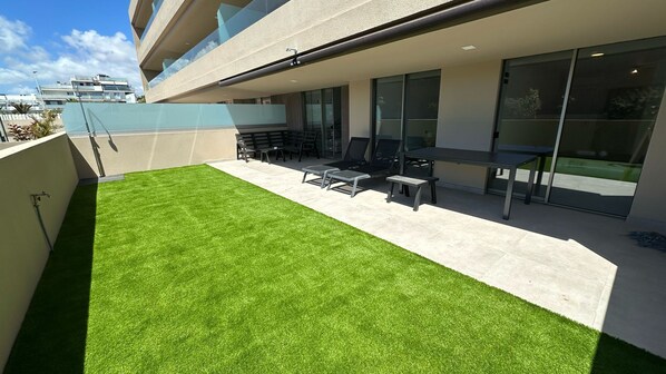 A modern outdoor patio with artificial grass, a seating area with chairs and tables, and a glass door leading to a building.