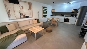 Una sala de estar moderna con un sofá, una mesa de centro y un reposapiés. Un comedor con mesa y sillas está adyacente a la cocina, que cuenta con microondas, horno y refrigerador.