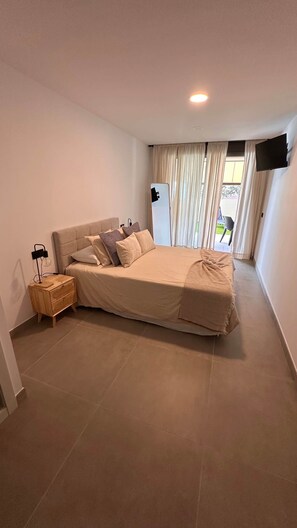 Un dormitorio con una cama, una mesita de noche, un televisor montado en la pared y un ventanal con cortinas.