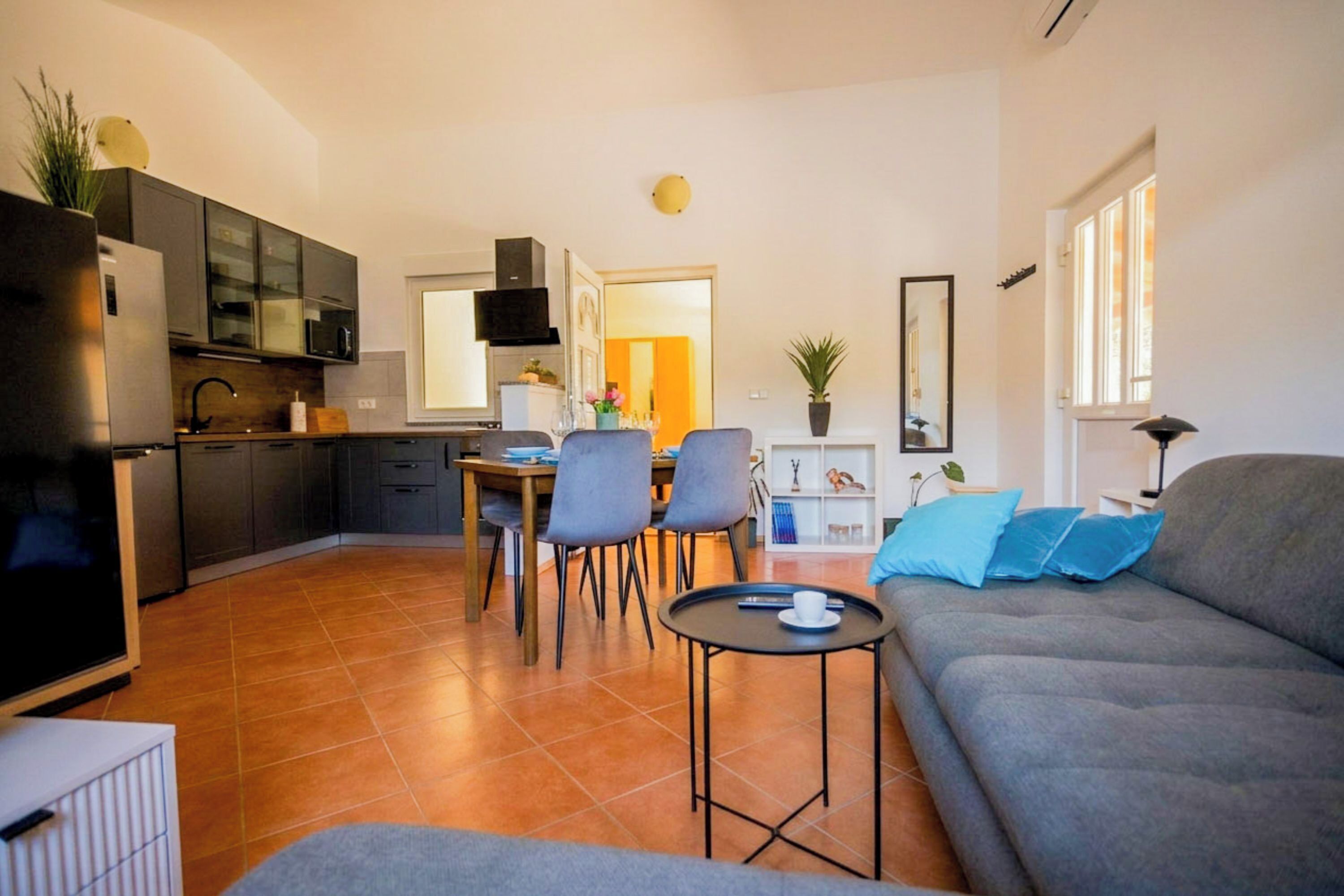 Een moderne woonkamer met een grijze bank, een eethoek met stoelen, een keuken met donkere kastjes en een zichtbaar raam.