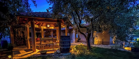 Un área de estar rústica al aire libre con una pérgola de madera, un barril y una mesa de piedra.