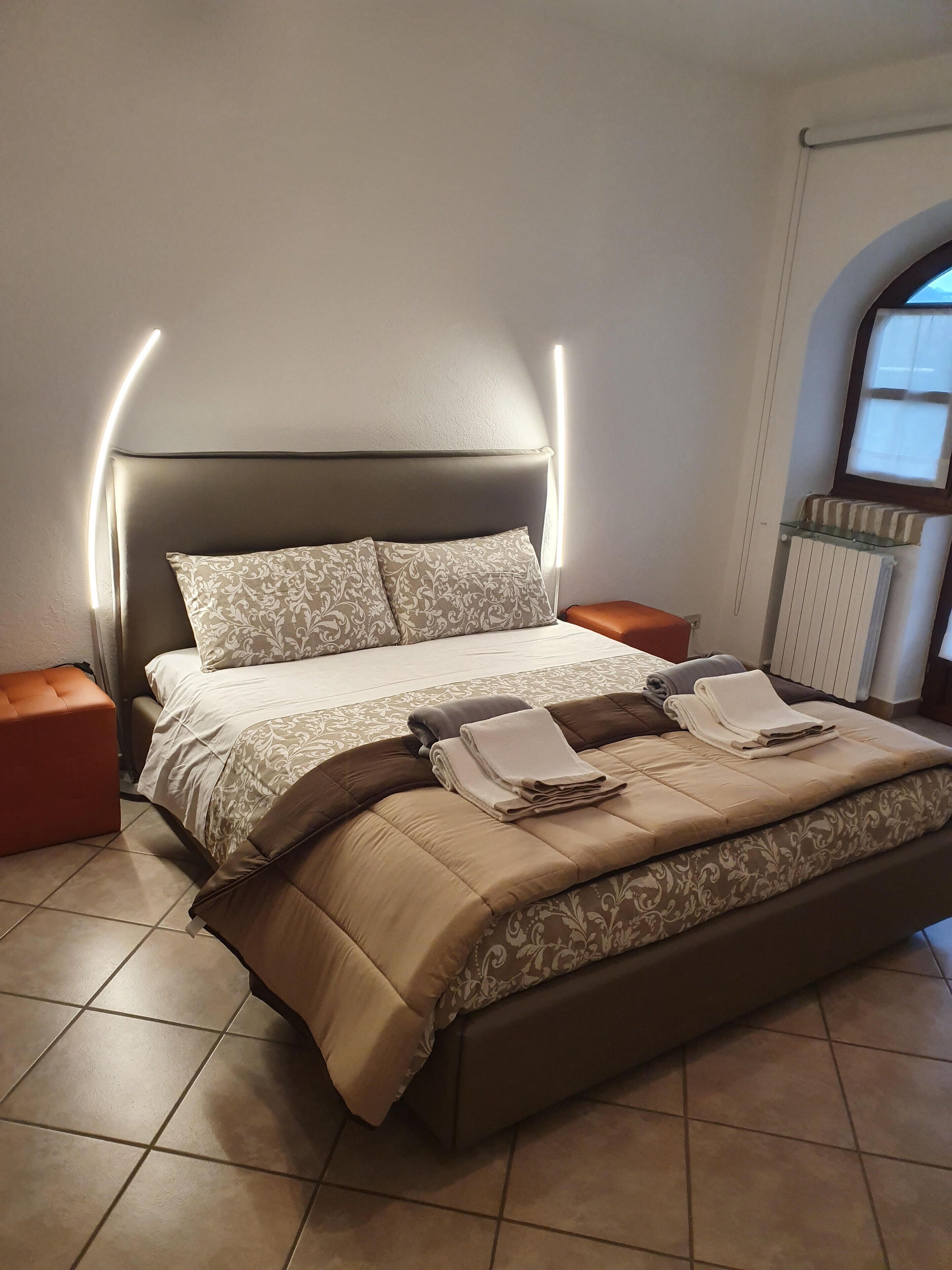 A neatly made bed with a patterned bedspread and pillows, a bedside table with a lamp, and a window with a view of the outside.