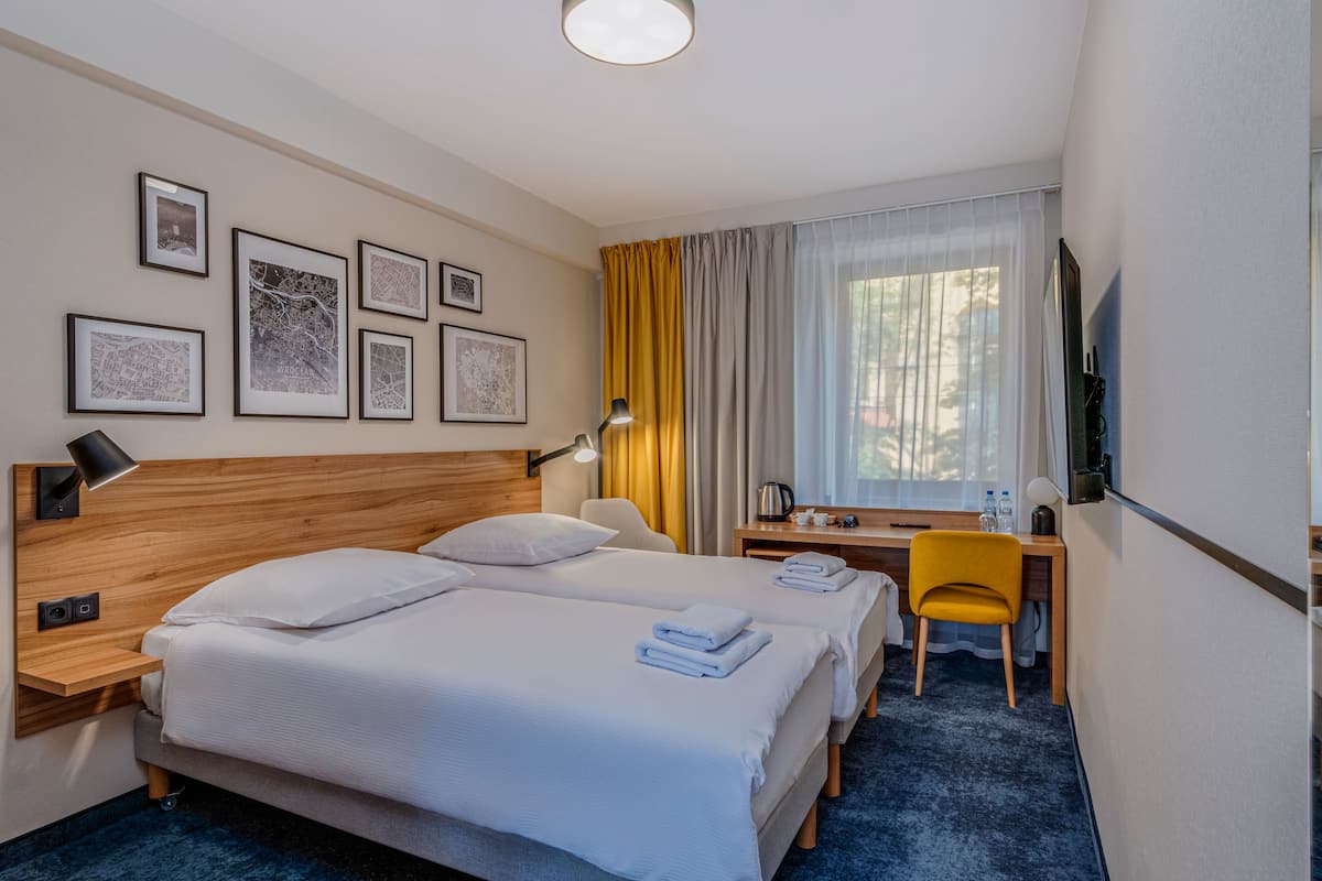 A hotel room with two beds, a desk, a chair, a TV, and artwork on the wall.