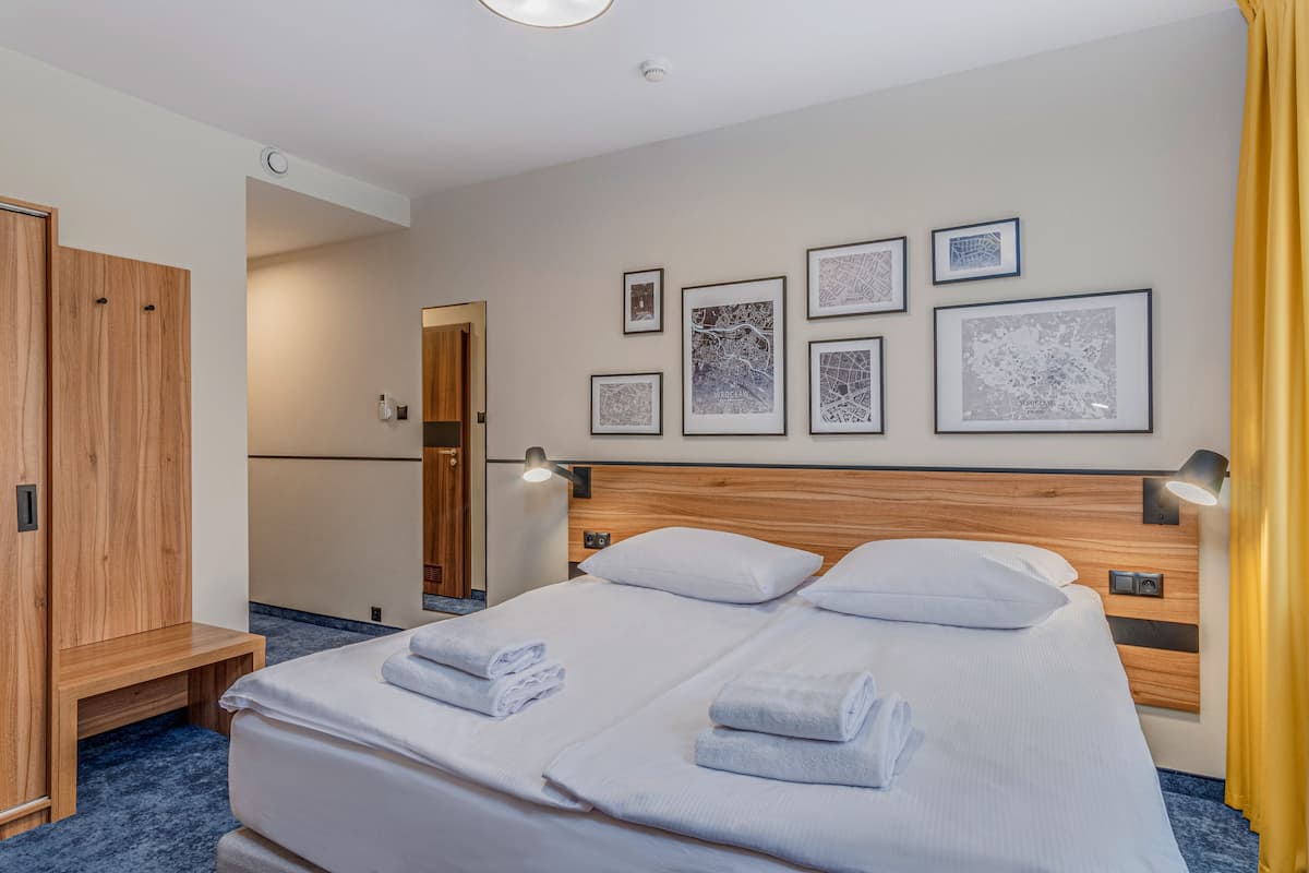 A hotel room with a large bed, two bedside tables, a wooden wardrobe, and a wall with framed maps.