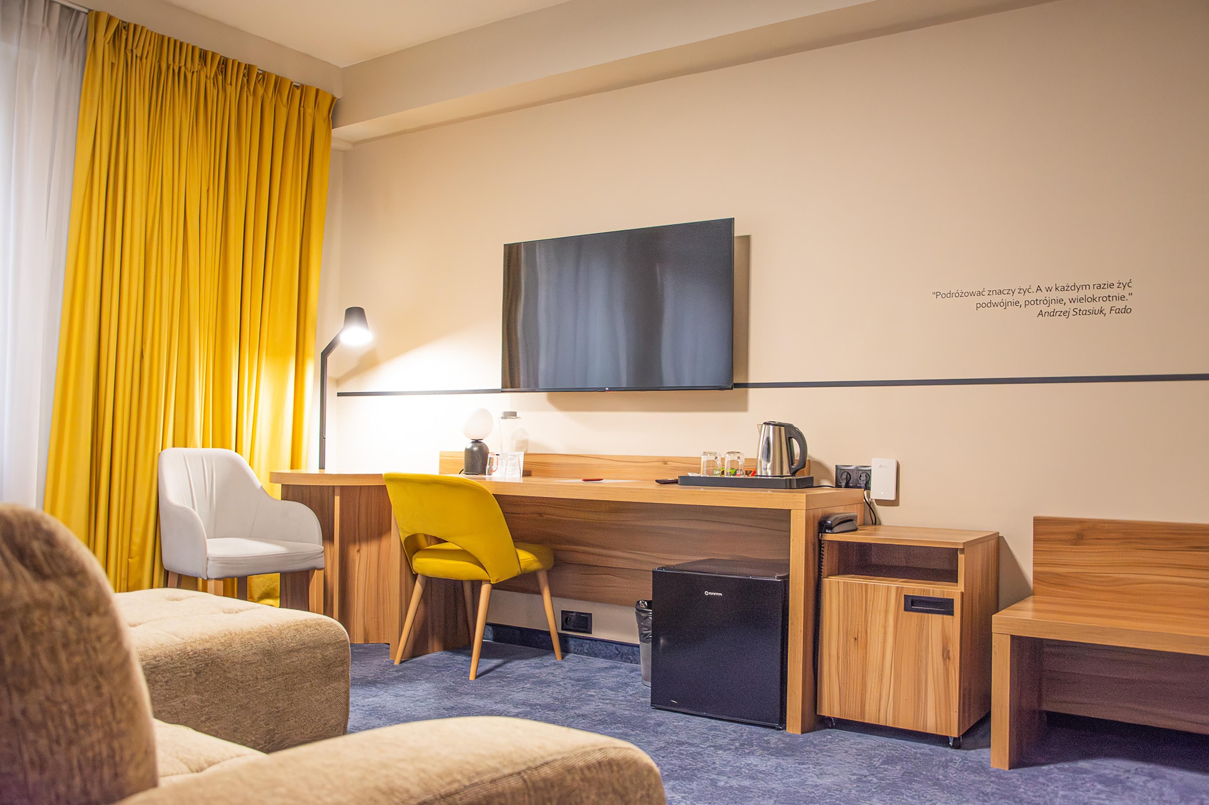 Una habitación de hotel con un escritorio de madera, una silla amarilla, una silla blanca, un cesto de basura negro pequeño y un televisor de pantalla plana montado en la pared.