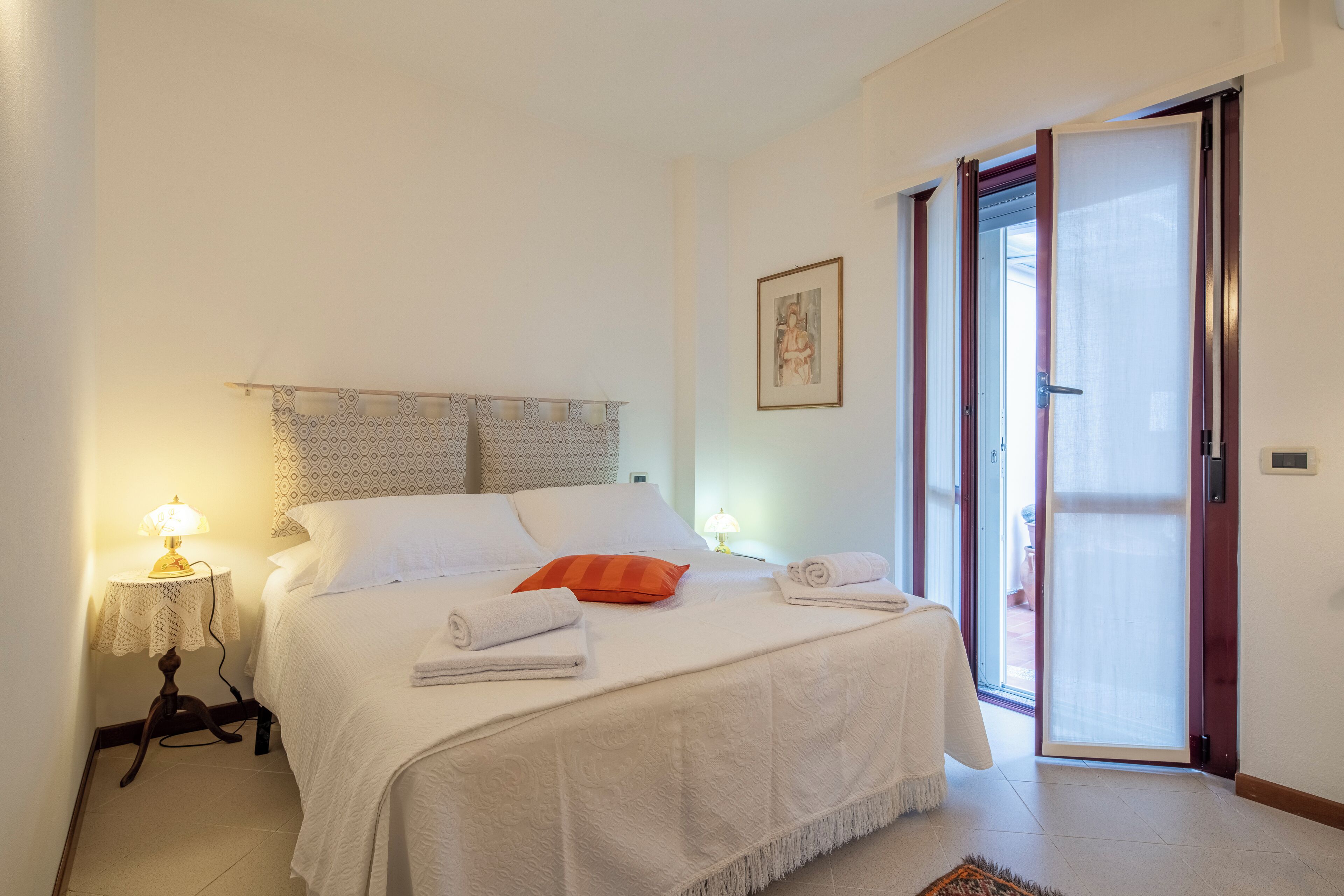 A neatly made bed with white linens and an orange pillow, a bedside lamp, and a framed picture on the wall.
