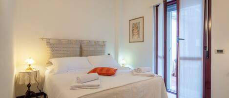 A neatly made bed with white linens and an orange pillow, a bedside lamp, and a framed picture on the wall.