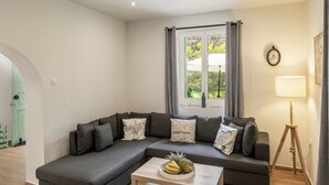 Una sala moderna con un sofá seccional gris, una mesa de centro de madera con fruta, una lámpara de pie y un ventanal con cortinas.