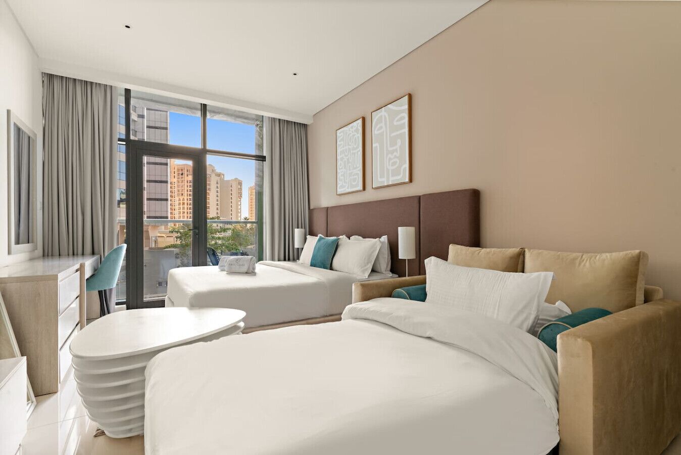 Une chambre d’hôtel avec un grand lit, un canapé, un bureau et une chaise. Il y a une fenêtre avec des rideaux, et deux tableaux encadrés accrochés au mur.