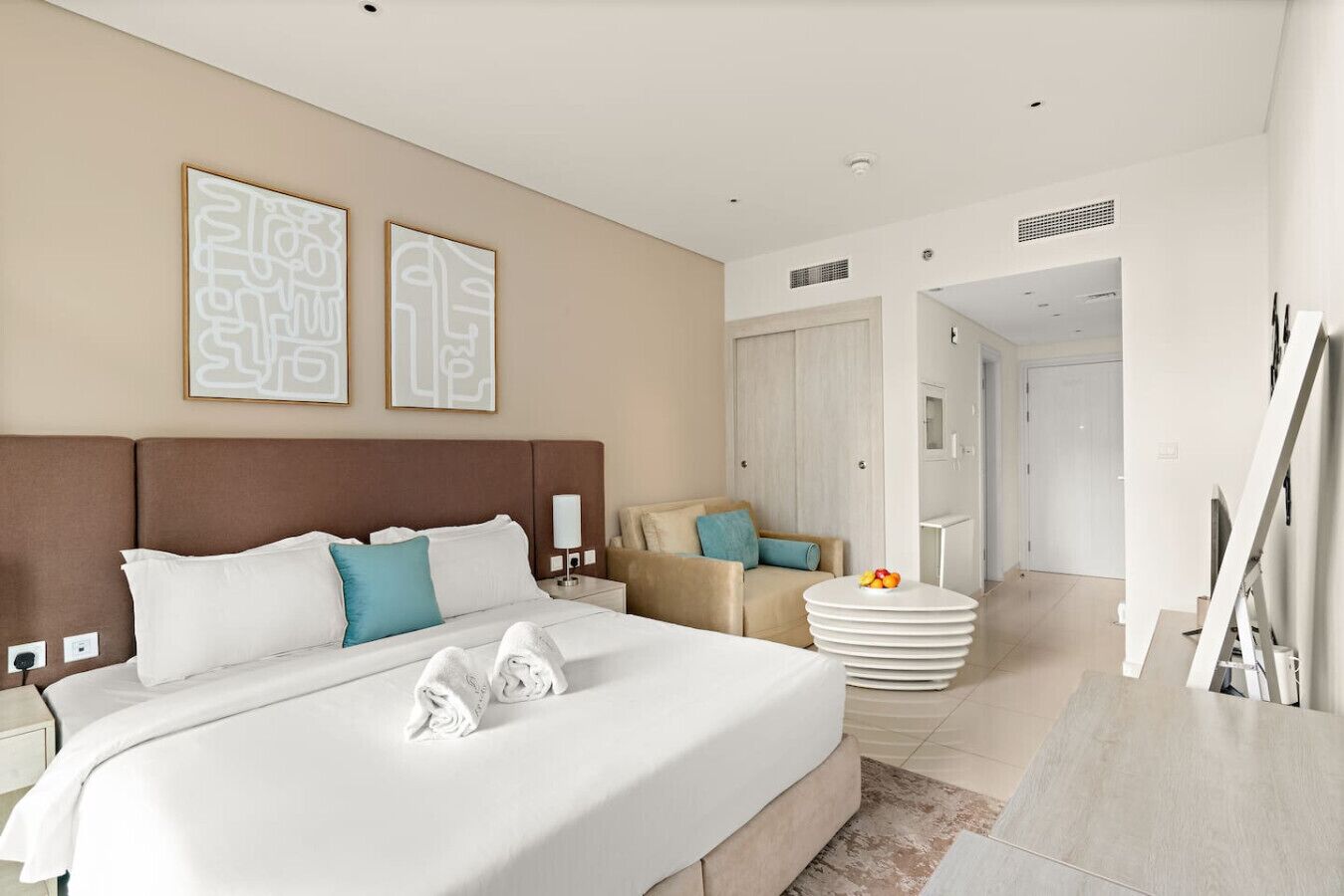 Une chambre d’hôtel moderne avec un grand lit, une table de chevet, une chaise et une petite table ronde sur laquelle sont disposés des fruits.