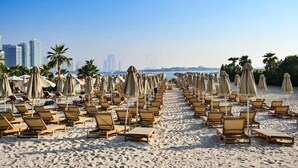 A beach with rows of sun loungers and umbrellas, palm trees, and a cityscape in the background. - FIRST CLASS | Studio | Palm Paradise (Dubai)