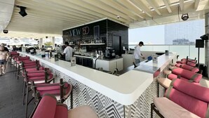 A rooftop bar with a long counter, red bar stools, and a bar area with a black countertop and shelves. - FIRST CLASS | Studio | Palm Paradise (Dubai)