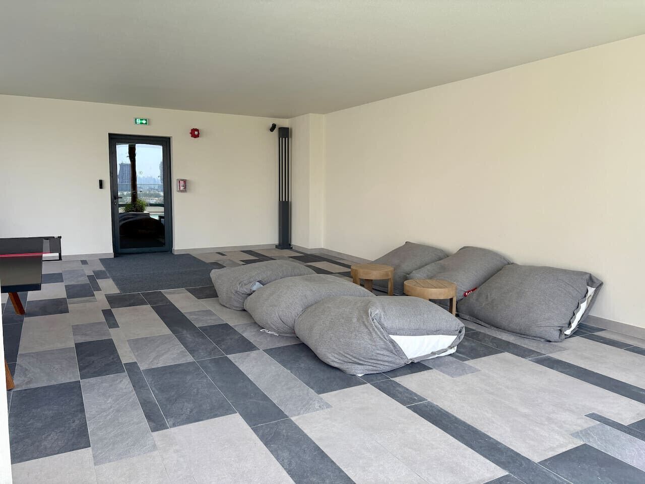 A hotel room with a checkered floor, a bed with grey pillows, a small wooden table, and a door with an emergency exit sign.