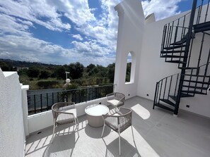 A balcony with a round table and two wicker chairs, a spiral staircase, and a view of greenery and buildings in the background. - Modern 3 bed townhouse with roof terrace, sleeps 6, in Aloha nr Puerto Banus! (Marbella)