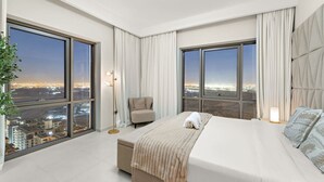 A modern hotel room with a large bed, a chair, and a floor lamp. There are large windows offering a city view.