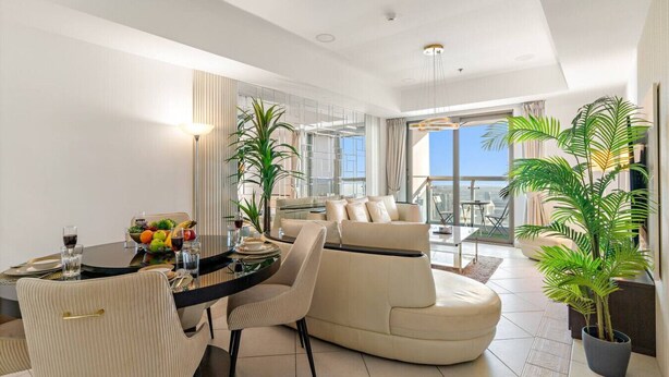 A modern dining area with a round table, chairs, and a fruit bowl. There is a balcony with a view of the city.