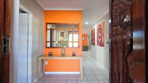 A hallway with an orange wall, a wooden door with an elephant carving, and a mirror above a console table with decorative vases. - Sunset Events and Accommodation  (Gqeberha)