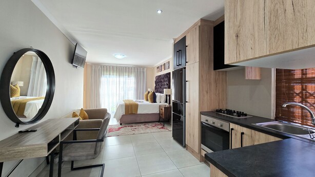 A modern hotel room with a bed, a sitting area with a table and chairs, a kitchen with a stove and sink, and a large mirror. - Sunset Events and Accommodation  (Gqeberha)