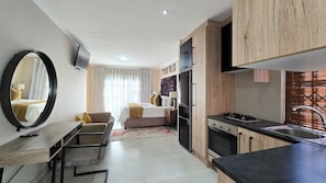 A modern hotel room with a bed, a sitting area with a table and chairs, a kitchen with a stove and sink, and a large mirror. - Sunset Events and Accommodation  (Gqeberha)