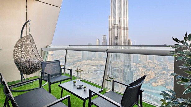 A balcony with a hanging chair, a table with two chairs, and a view of a tall skyscraper. - Burj Khalifa View | 4BR Home | Walk to Dubai Mall | Pool & Parking (Downtown)