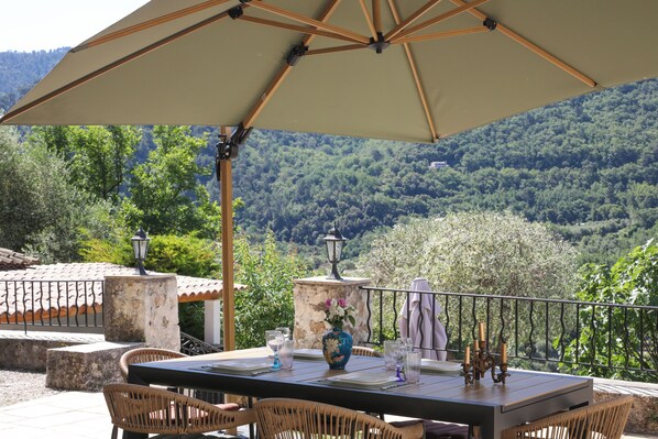 An outdoor dining area with a table set for two, surrounded by wicker chairs, under a large umbrella.