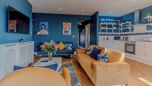 Een moderne woonkamer met een bank, salontafel en televisie. Er is een keuken met witte kasten en een neonbord.
