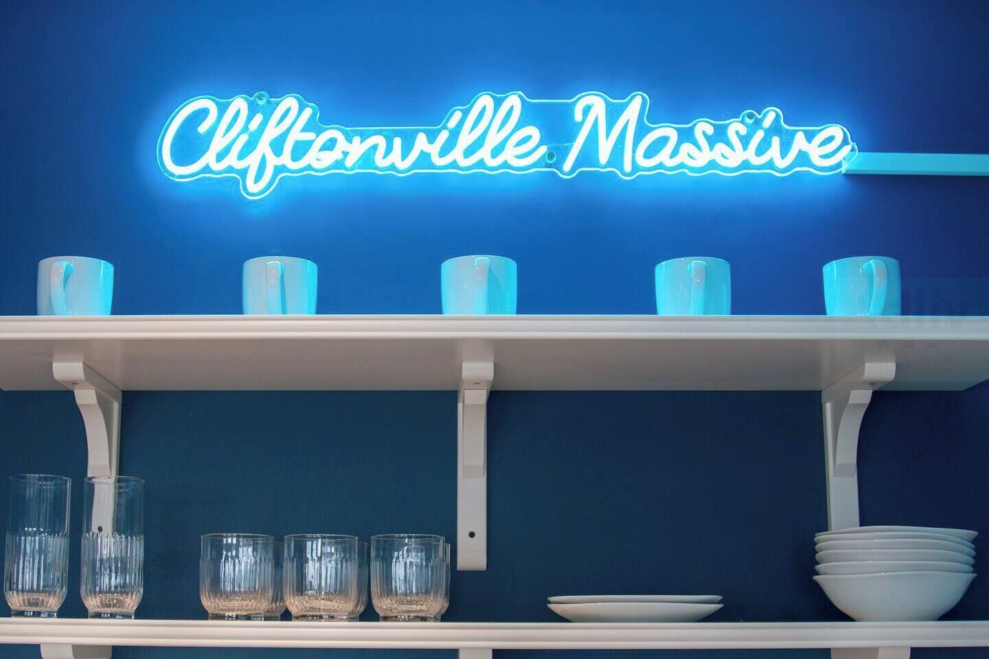 A neon sign reading "Cliftonville Massive" above a shelf with blue cups and glasses.