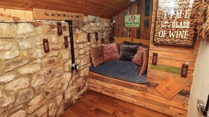 Une pièce chaleureuse avec un mur en pierre, un plafond en bois et un lit agrémenté d’oreillers. On peut y voir des panneaux décoratifs sur le mur, dont l’un porte l’inscription : « Ici, il y a toujours le temps de savourer un verre de vin. »