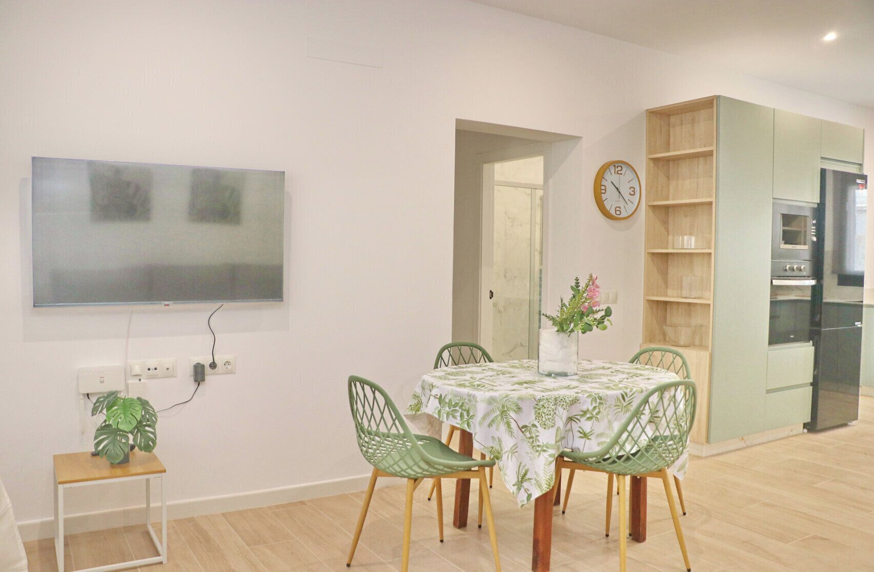 A modern kitchen with a built-in oven and refrigerator, a dining area with a table and chairs, and a wall-mounted clock.