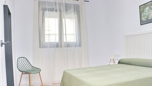 A bedroom with a bed, a chair, a window with sheer curtains, and a radiator.
