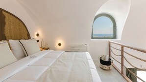 A bedroom with a large bed, a small bedside table with a lamp, a window with a view, and a balcony with a rope railing. - Acro Caldo Suites (Akrotiri)