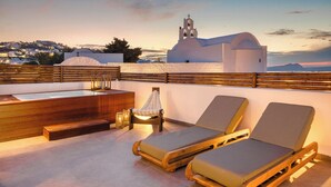 A rooftop terrace with two lounge chairs, a wooden bench, and a small table with a lamp. In the background, there is a white building with a bell tower and a dome. - Acro Caldo Suites (Akrotiri)