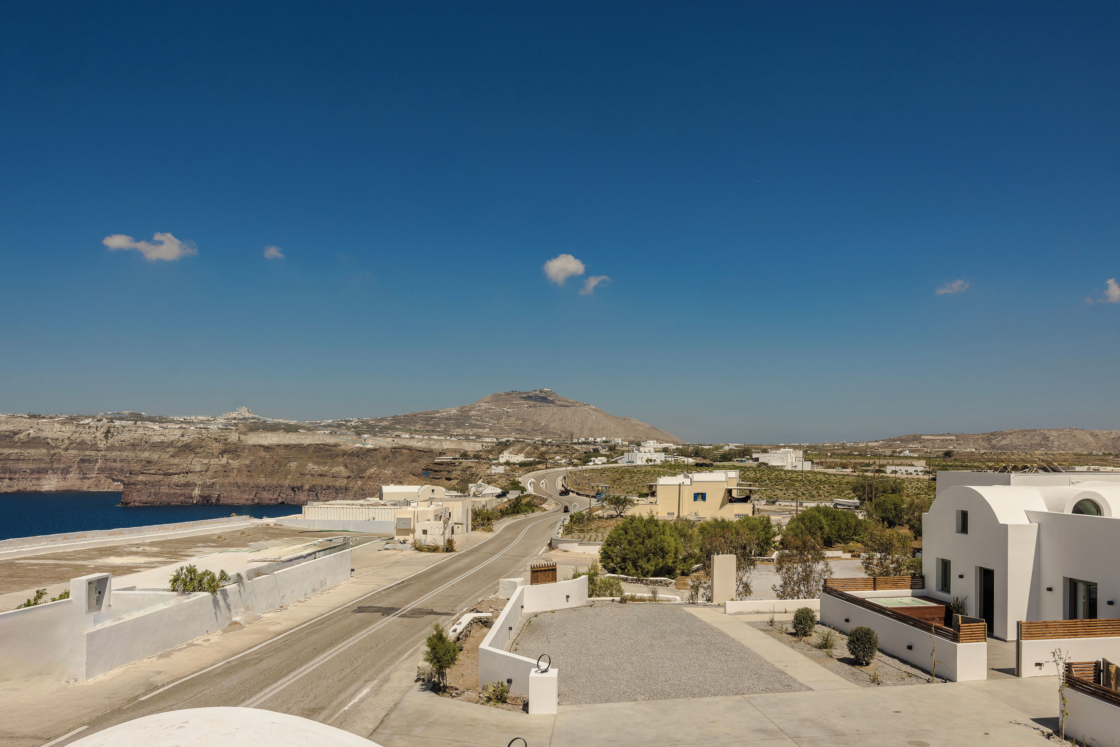 A coastal town with modern architecture, a clear blue sky, and a road leading towards a hilly landscape.
