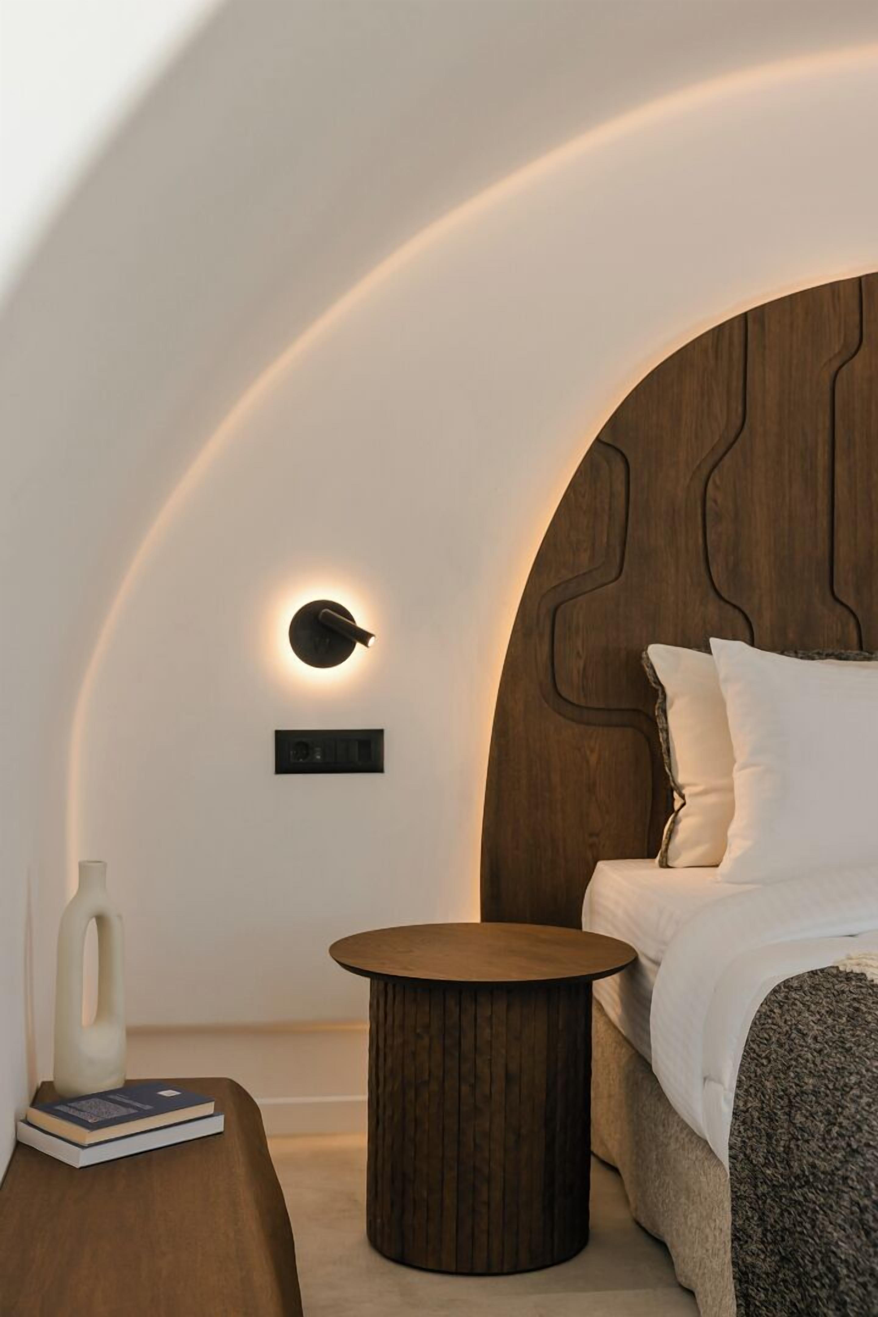 Un dormitorio moderno con techo curvo, cabecera de madera, mesita de noche y velador.