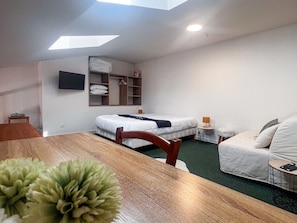 A hotel room with a wooden dining table, two beds, a sofa, a TV mounted on the wall, and a skylight.