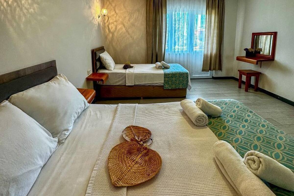 A hotel room with two beds, a large window with curtains, a small table with a mirror, and a wall-mounted light fixture.