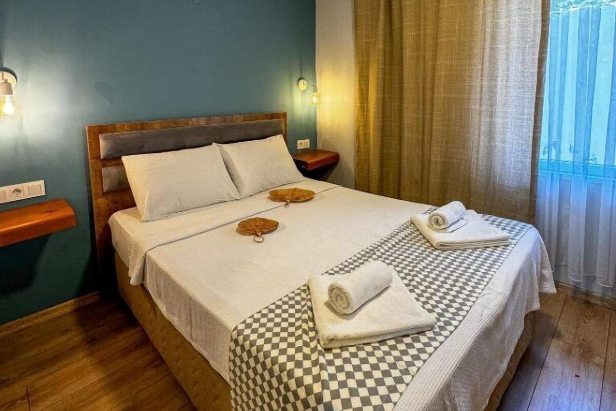 A hotel room with a large bed, wooden headboard, two bedside tables, and a view of the outdoors through a window with curtains.