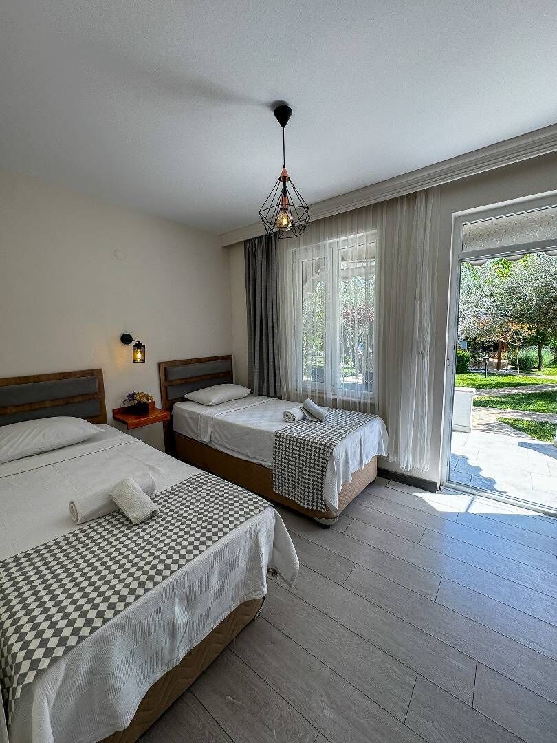 Habitación de hotel con dos camas, un ventanal con cortinas, una mesita con lámpara y vistas al exterior.