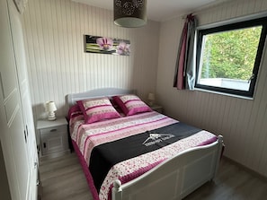 Une chambre à coucher comprenant un lit, une table de chevet, une lampe, une fenêtre avec des rideaux et un tableau accroché au mur.
