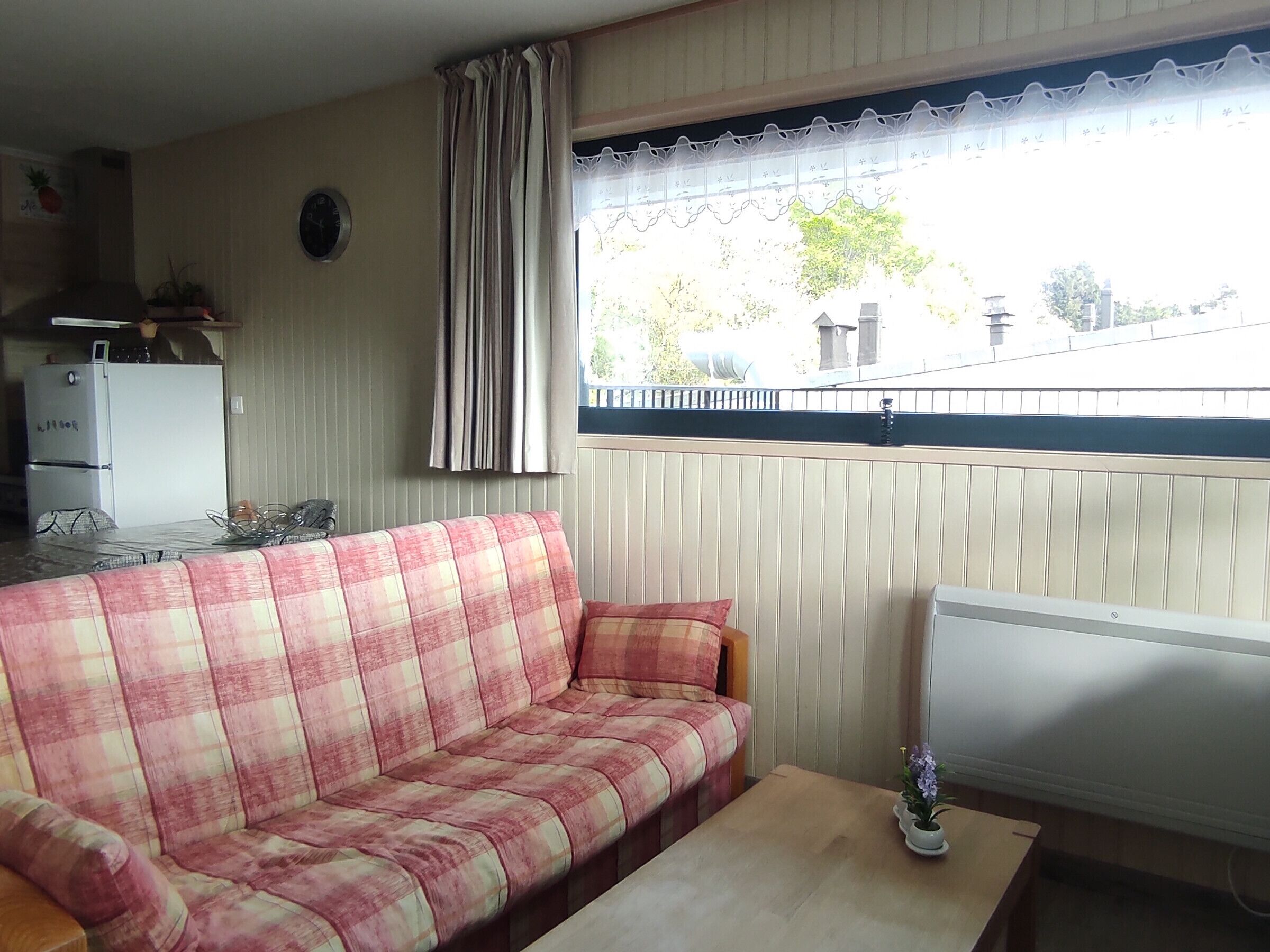 Ett vardagsrum med en soffa, ett litet bord, ett element, ett fönster med gardiner, en klocka och en köksdel med ett kylskåp och en spis.
