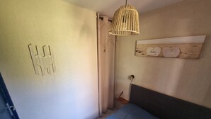Une chambre avec un lit, un luminaire suspendu, une étagère fixée au mur avec des objets décoratifs, et une porte dotée d’un porte-serviettes.